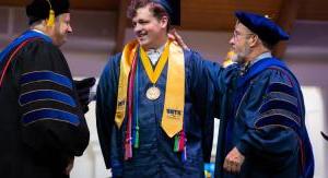 Two professors in academic regalia place a stole around a male graduate on a stage.