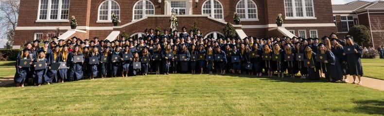Large group of graduates posed with diplomas in front of brick academic building smiling at the camera