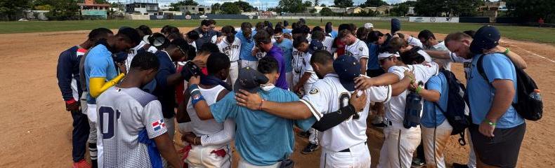 Baseball players praying in a circle with their arms around each other outdoors on a baseball field
